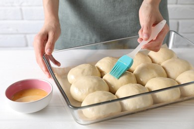 Photo of Woman spreading egg yolk onto raw dough balls at white wooden table, closeup