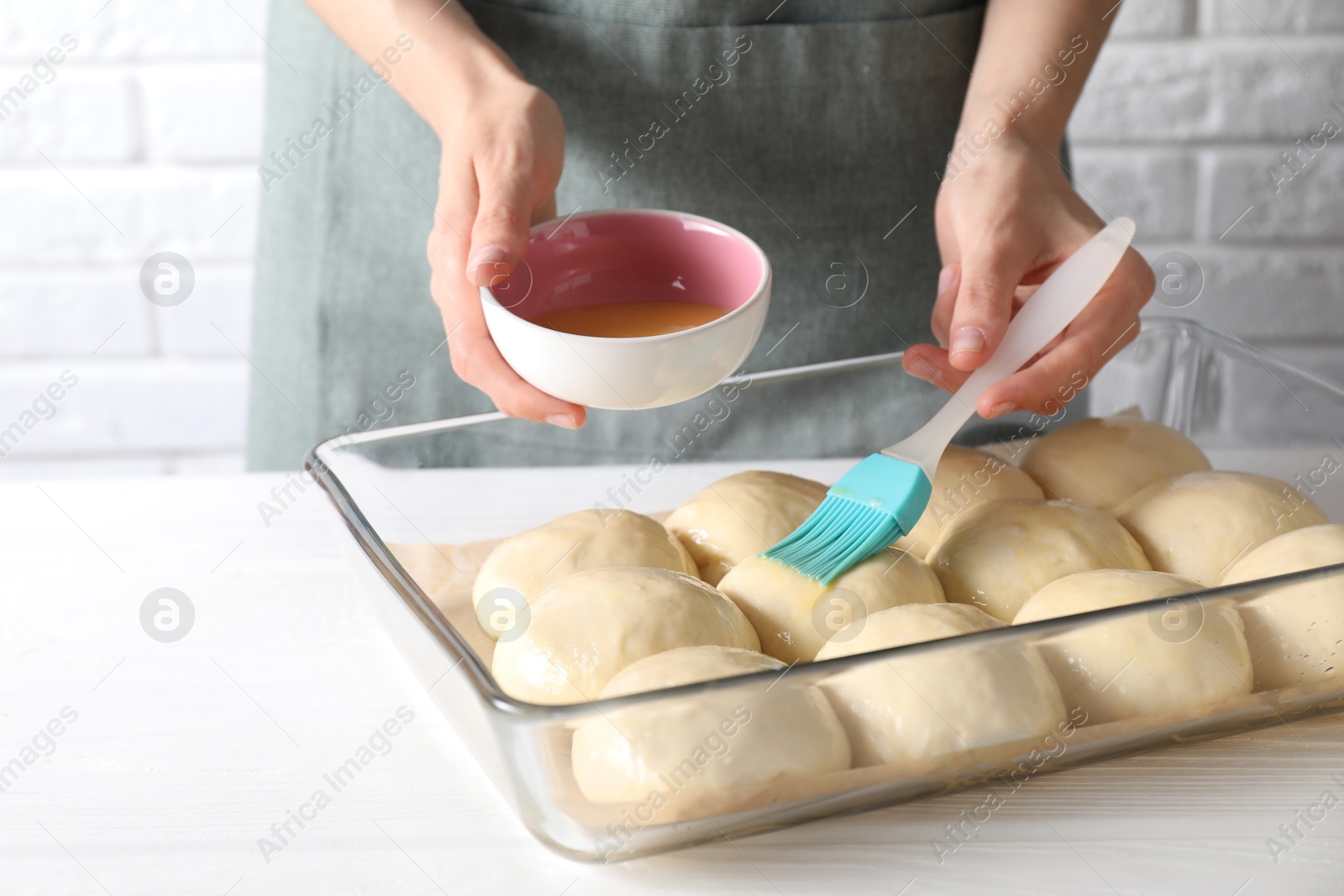 Photo of Woman spreading egg yolk onto raw dough balls at white wooden table, closeup