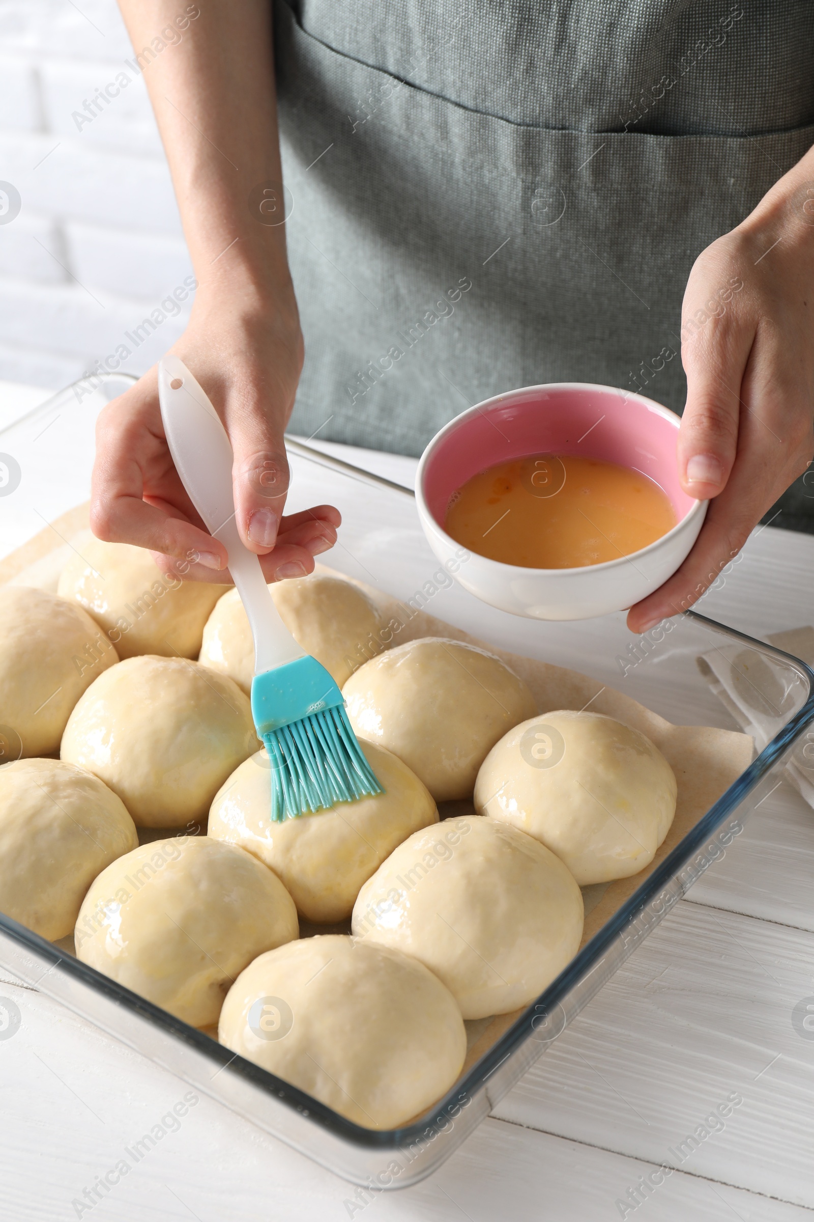 Photo of Woman spreading egg yolk onto raw dough balls at white wooden table, closeup