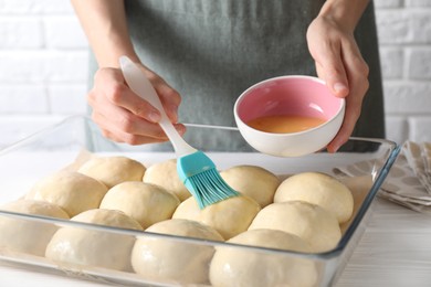 Woman spreading egg yolk onto raw dough balls at white wooden table, closeup