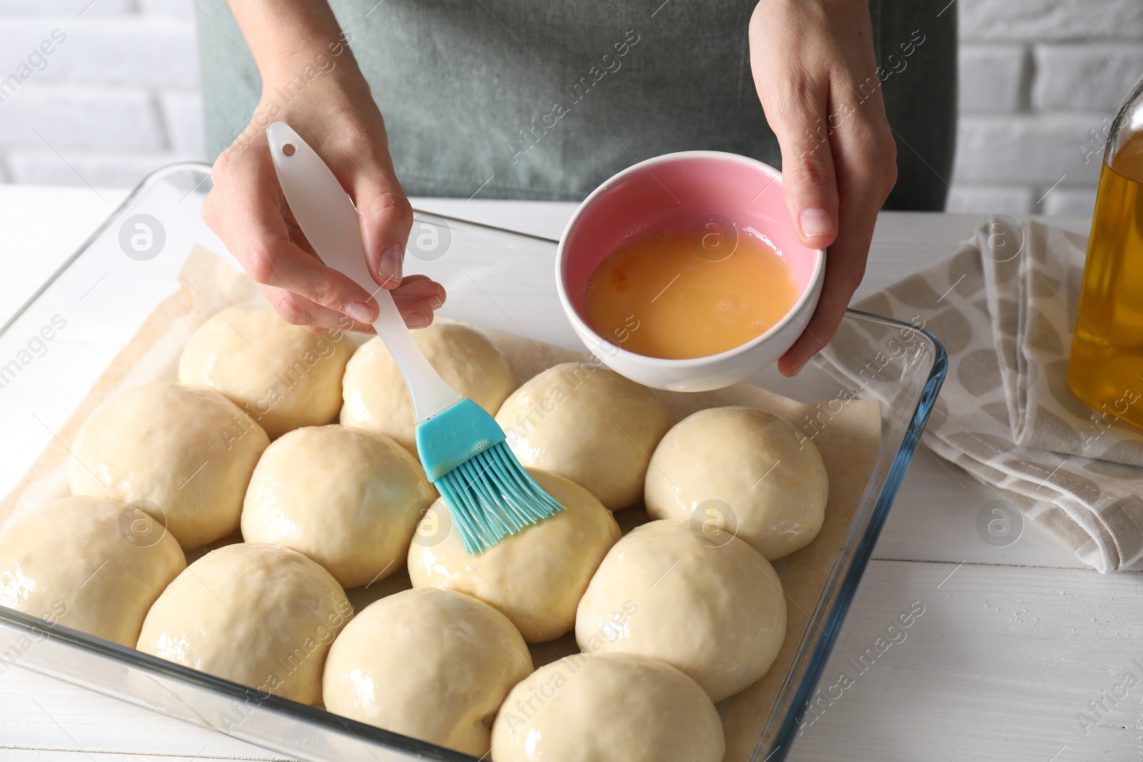 Photo of Woman spreading egg yolk onto raw dough balls at white wooden table, closeup