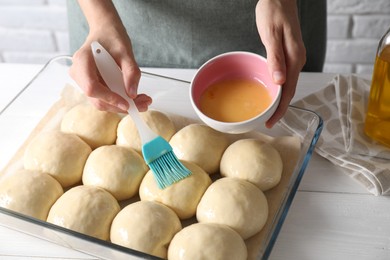 Woman spreading egg yolk onto raw dough balls at white wooden table, closeup