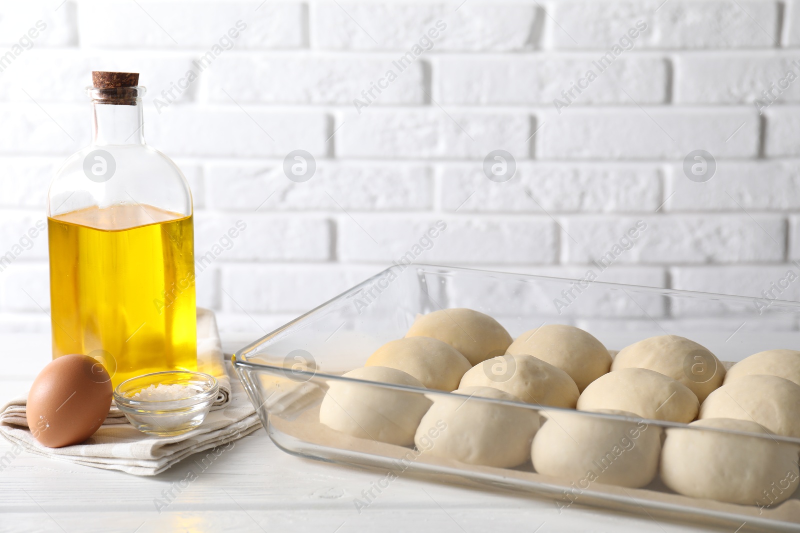 Photo of Raw dough balls in baking dish, egg, salt and oil on white wooden table