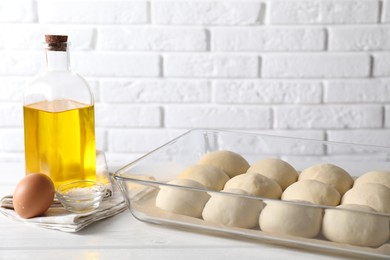 Raw dough balls in baking dish, egg, salt and oil on white wooden table