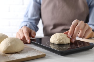 Photo of Woman weighing raw dough ball on kitchen scale at white wooden table, closeup