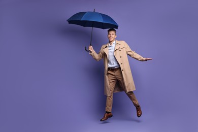 Photo of Young man with blue umbrella jumping on purple background, space for text