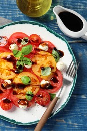 Photo of Tasty salad with balsamic vinegar, mozzarella and fork on blue wooden table, flat lay