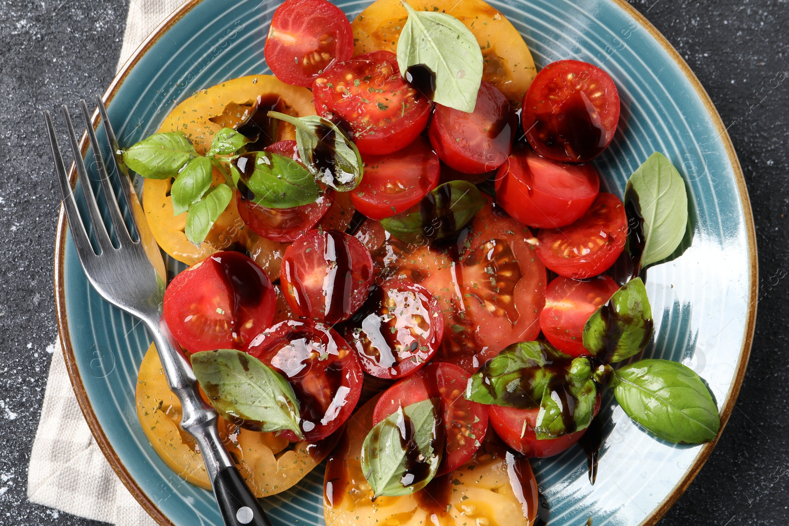 Photo of Tasty salad with balsamic vinegar, basil and fork on black textured table, top view