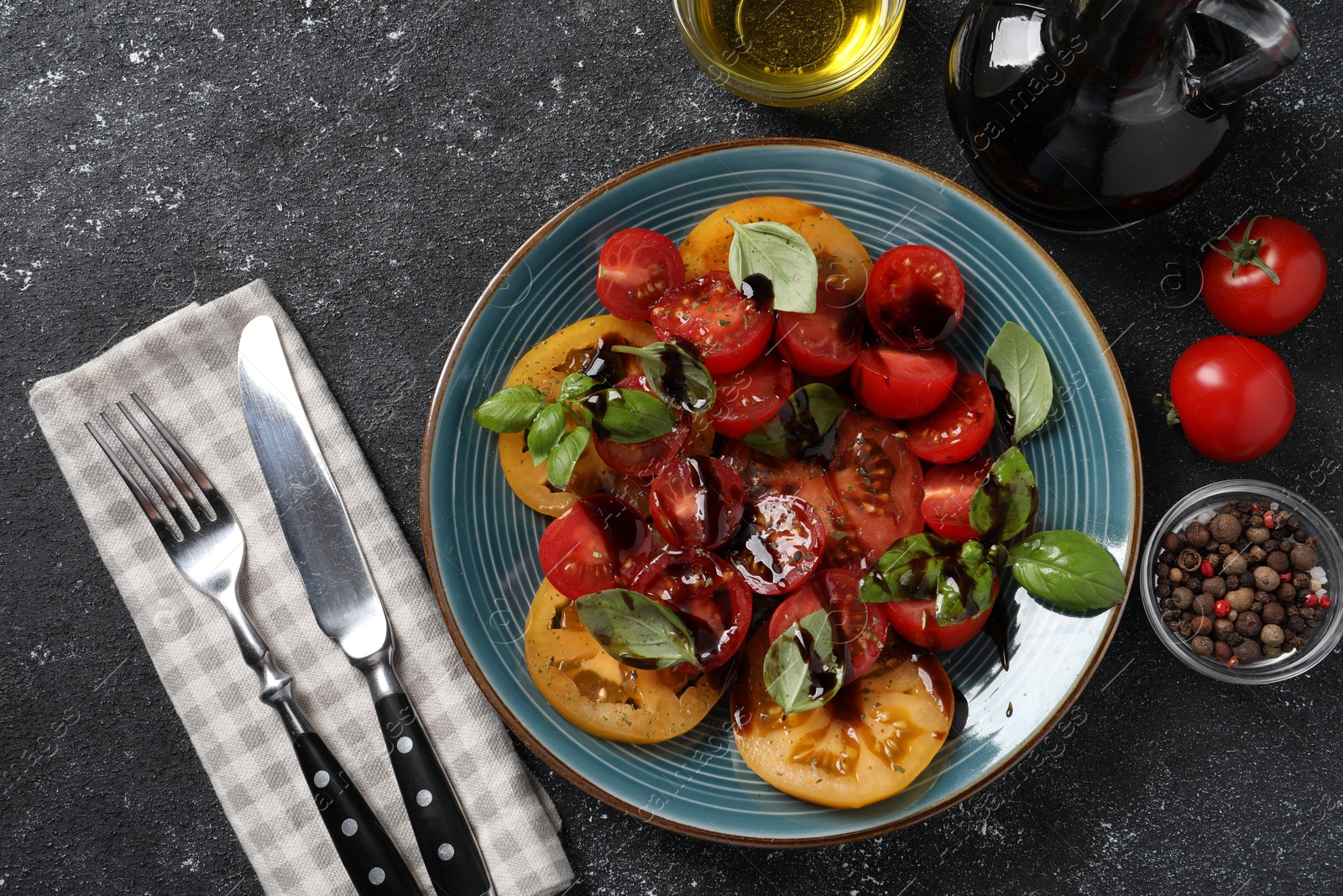 Photo of Tasty salad with balsamic vinegar, basil and cutlery on black textured table, flat lay