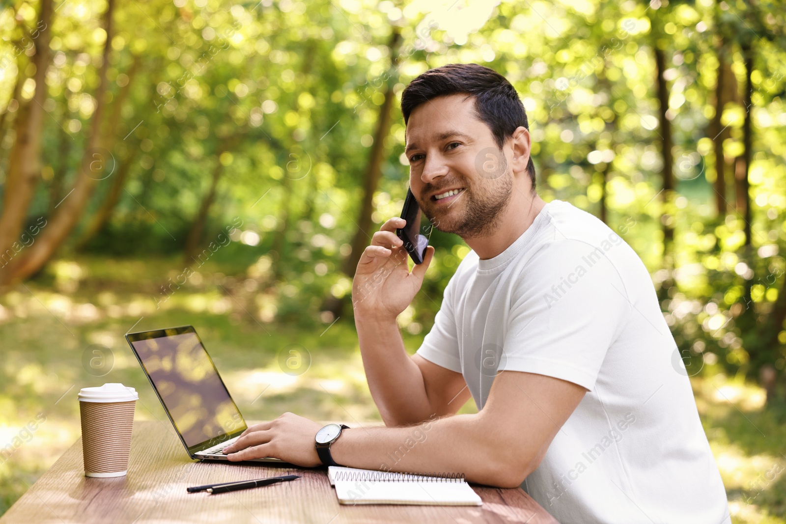 Photo of Smiling freelancer talking on smartphone at table with laptop outdoors. Remote job