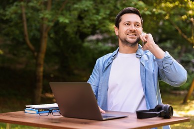 Photo of Smiling freelancer talking by smartphone at table with laptop outdoors, low angle view. Remote job