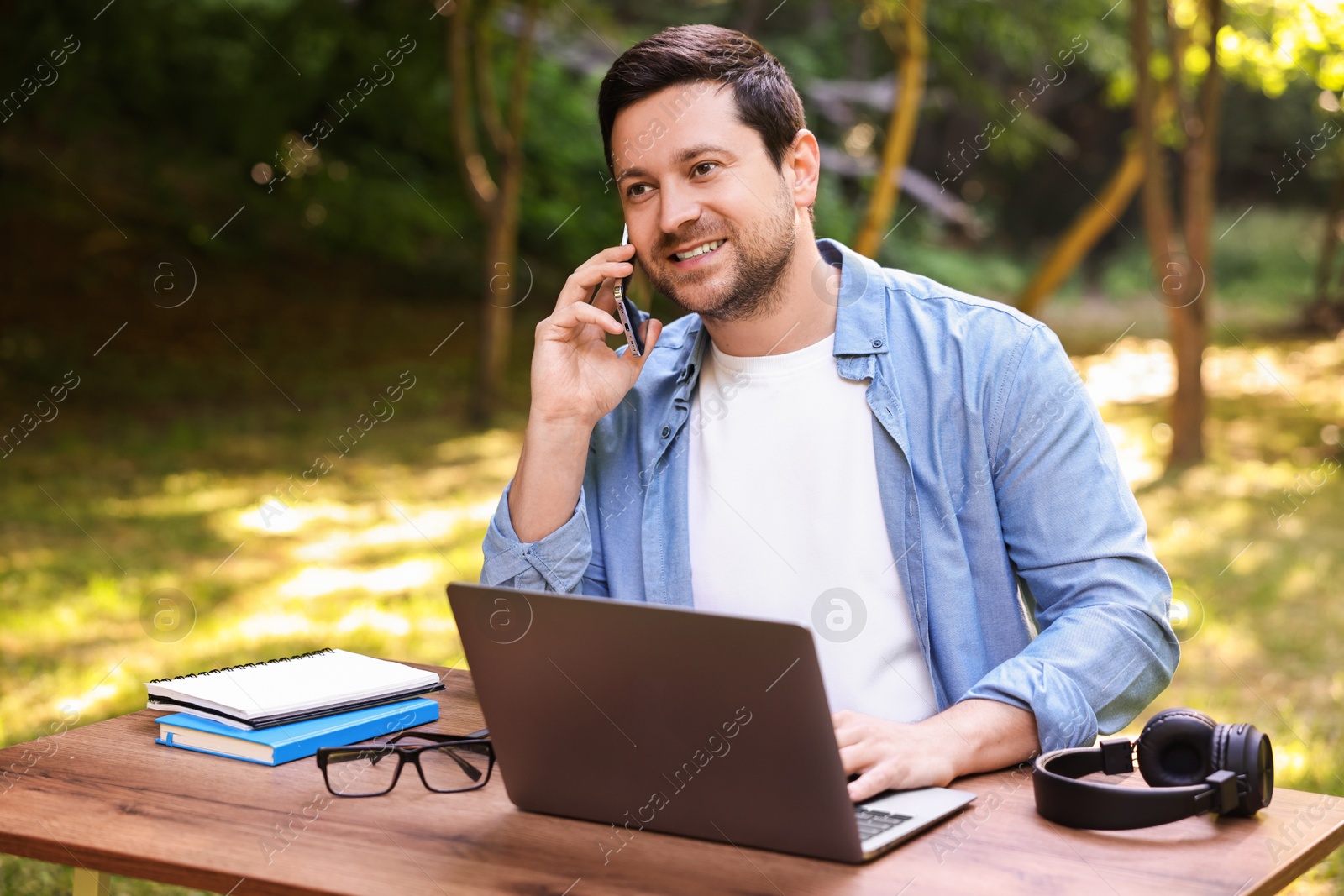 Photo of Smiling freelancer talking on smartphone at table with laptop outdoors. Remote job