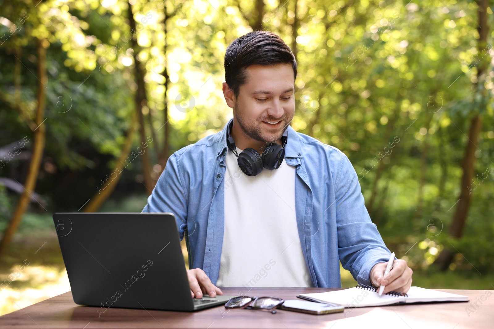 Photo of Smiling freelancer working with laptop and writing something at table outdoors. Remote job