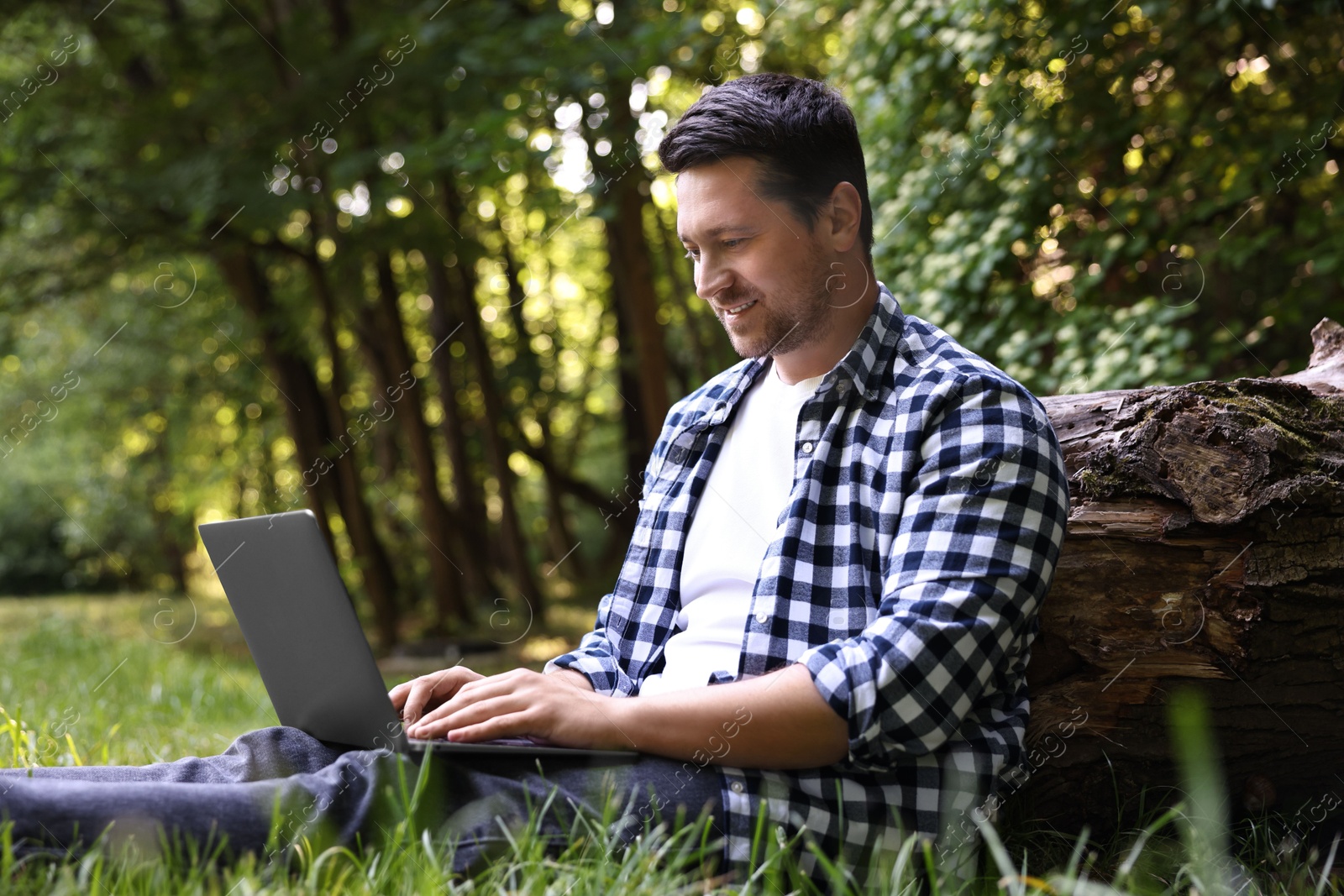 Photo of Smiling freelancer working with laptop on green grass in forest. Remote job