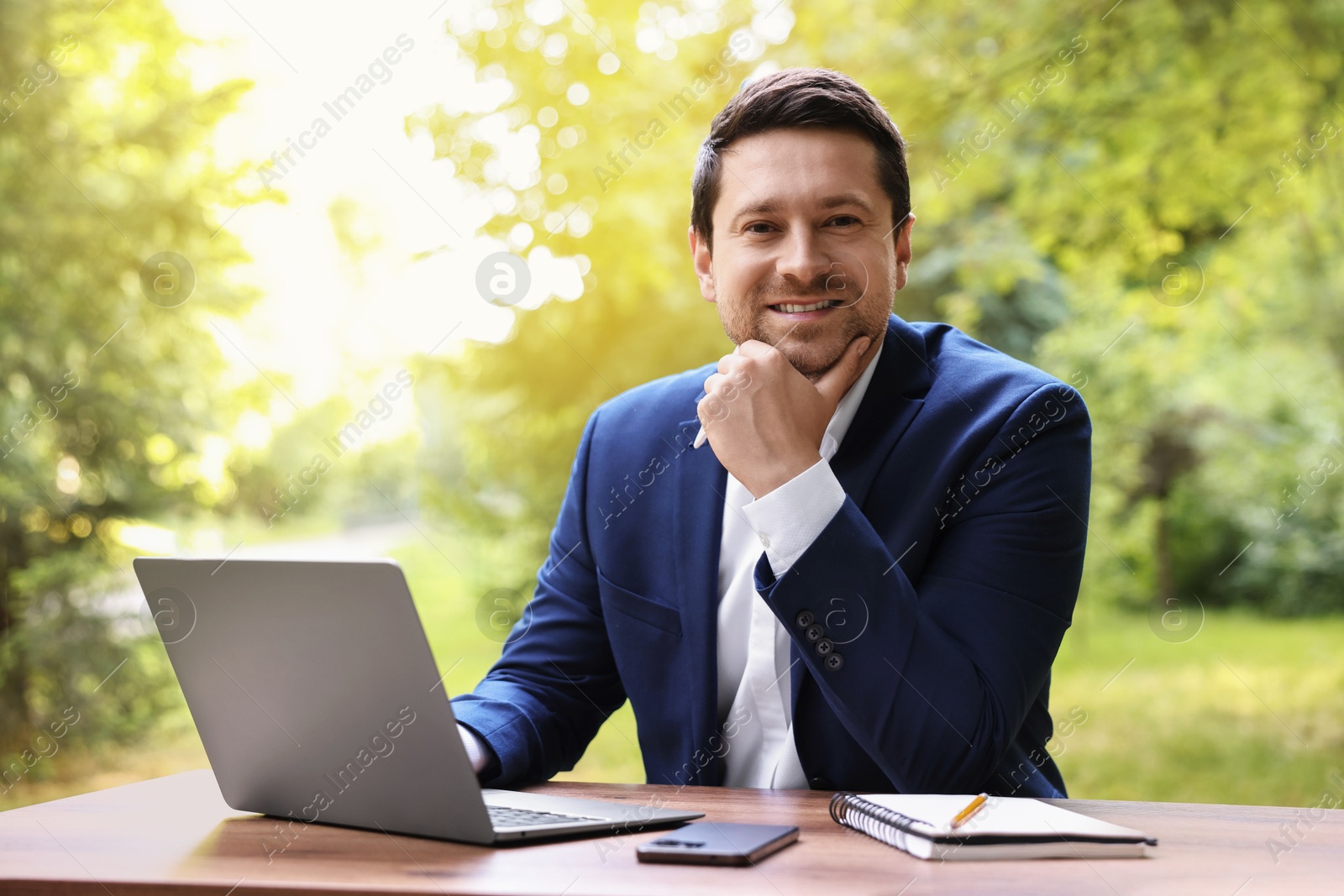 Photo of Smiling businessman working with laptop at table outdoors. Remote job