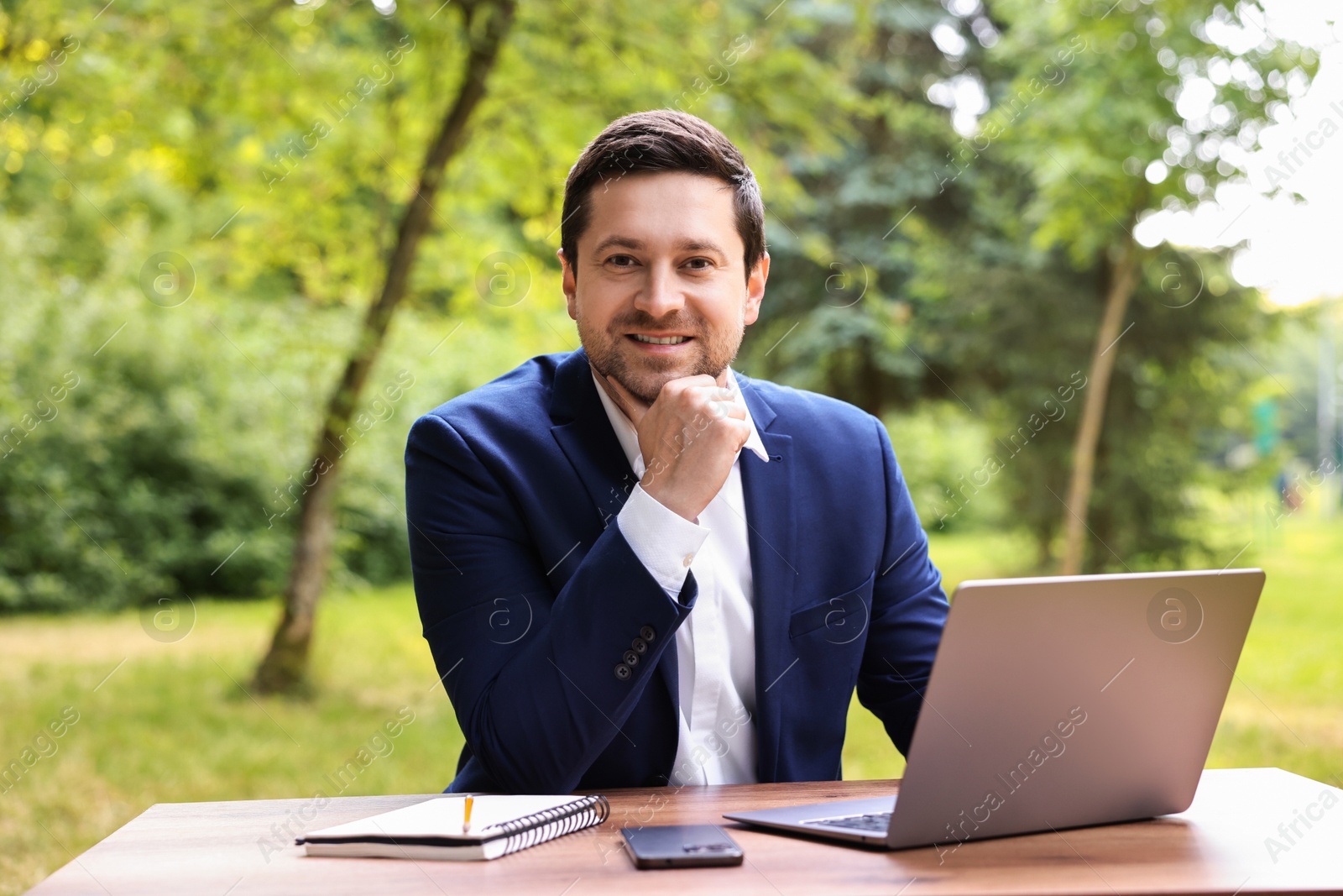 Photo of Smiling businessman working with laptop at table outdoors. Remote job