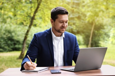 Photo of Smiling businessman working with laptop and writing something at table outdoors. Remote job