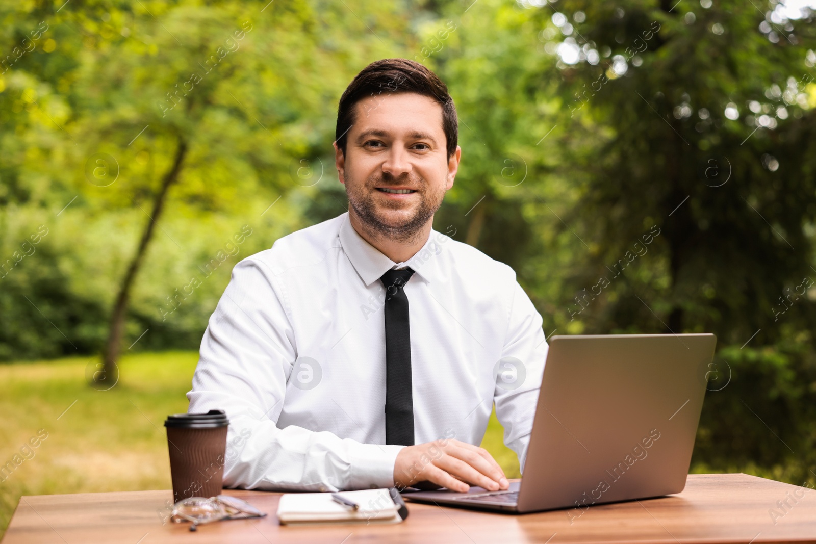 Photo of Smiling businessman working with laptop at table outdoors. Remote job