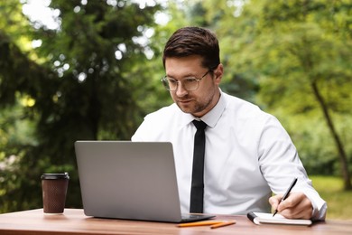 Photo of Businessman working with laptop and writing something at table outdoors. Remote job