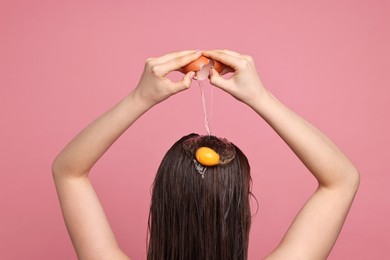 Photo of Woman breaking egg onto her head against pink background, back view. Natural hair mask