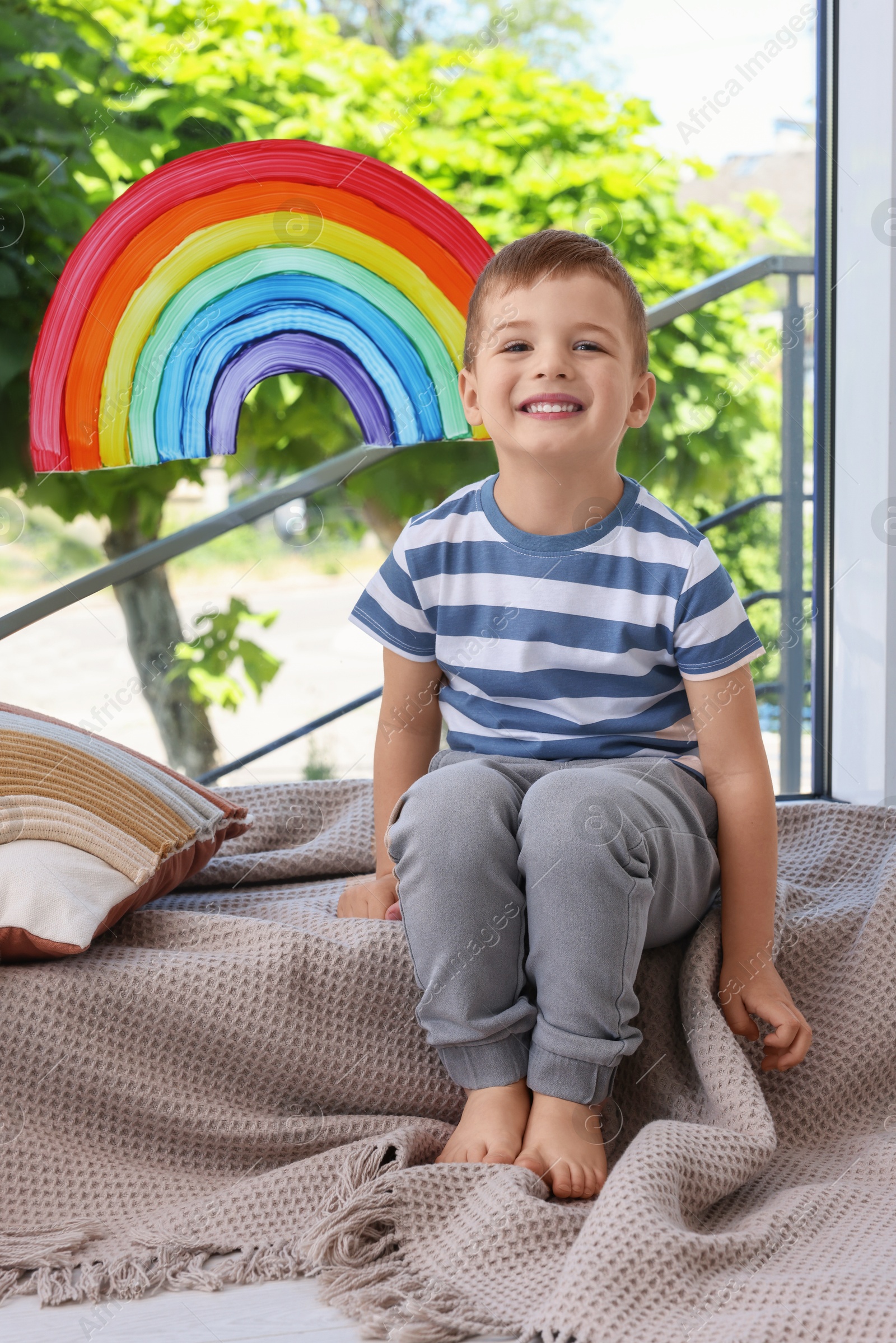 Photo of Happy little boy near rainbow painting on window indoors