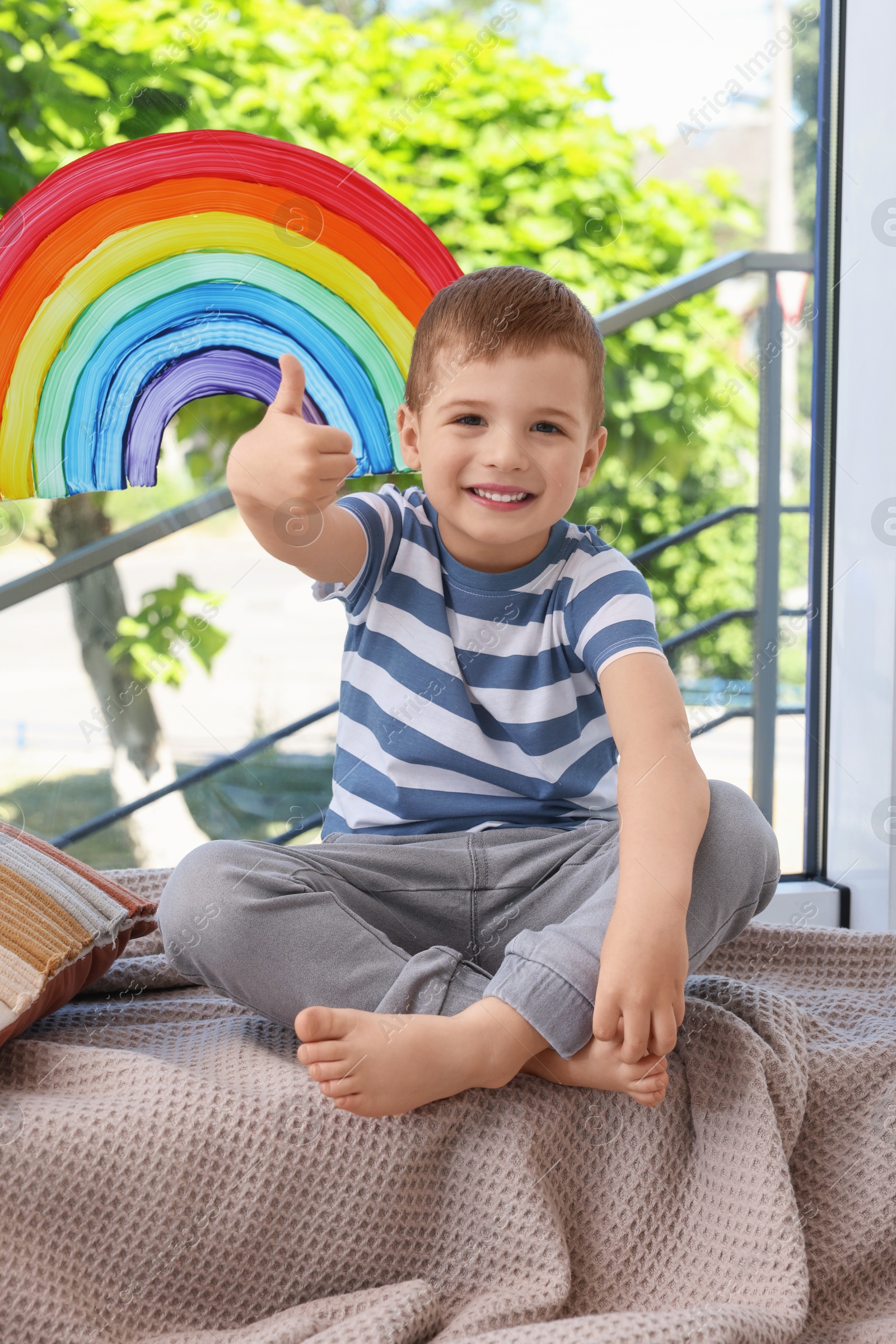 Photo of Happy little boy near rainbow painting on window indoors