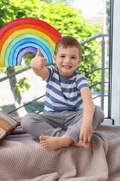 Photo of Happy little boy near rainbow painting on window indoors