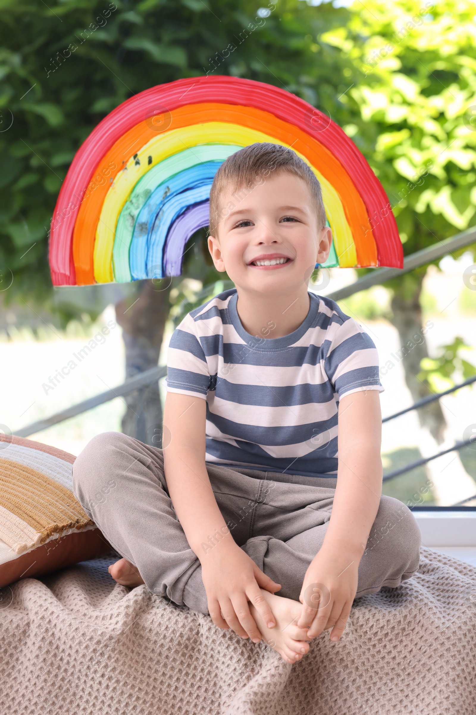 Photo of Happy little boy near rainbow painting on window indoors