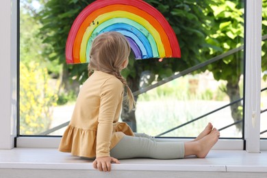 Photo of Little girl near rainbow painting on window indoors