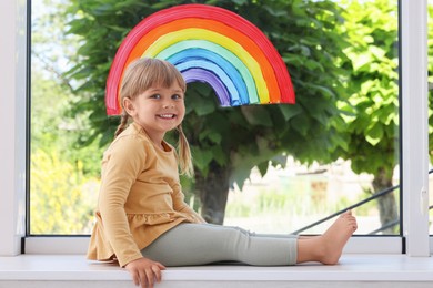 Little girl near rainbow painting on window indoors