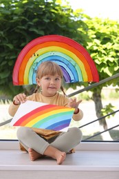 Little girl with picture of rainbow near window indoors