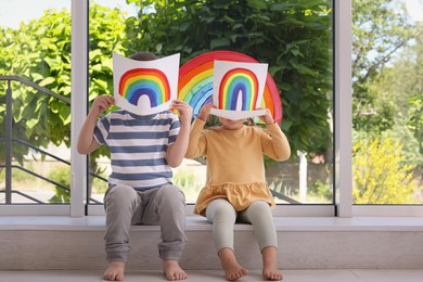 Children covering faces with pictures of rainbow near window indoors