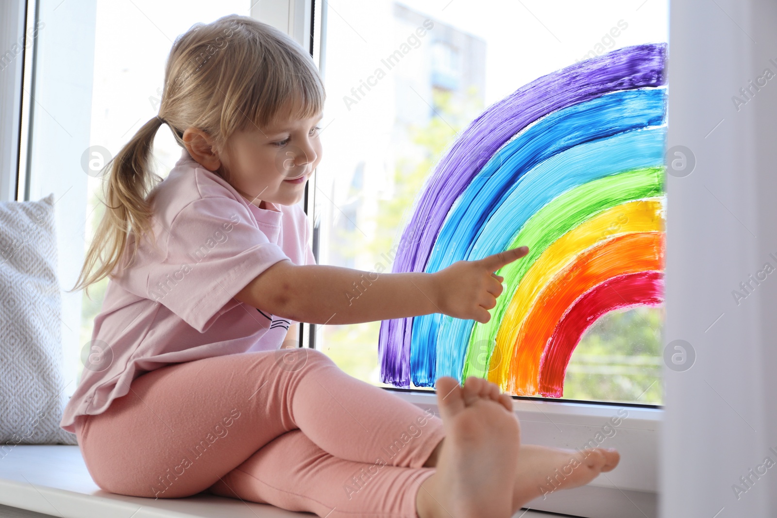 Photo of Little girl touching picture of rainbow on window indoors