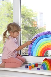 Photo of Little girl drawing rainbow on window indoors