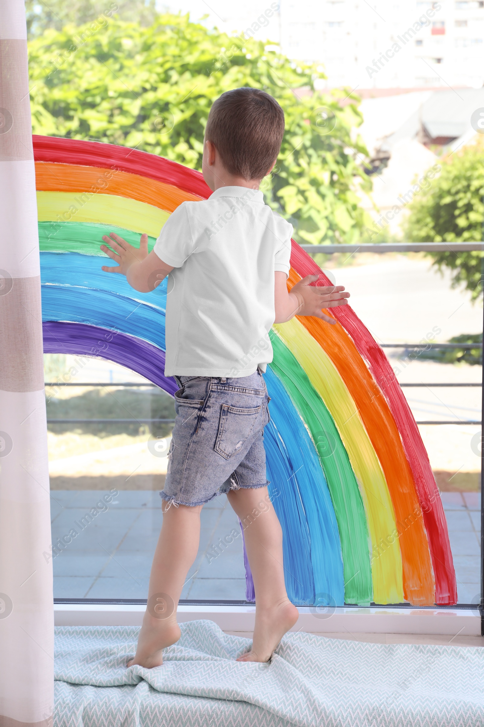 Photo of Little boy touching picture of rainbow on window indoors, back view