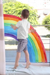 Photo of Little boy touching picture of rainbow on window indoors, back view