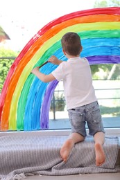 Photo of Little boy drawing rainbow on window indoors, back view