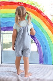 Photo of Little girl near rainbow painting on window indoors, back view