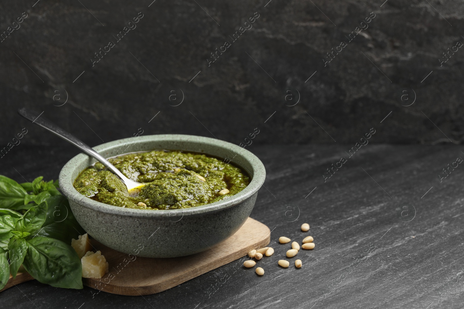 Photo of Tasty pesto sauce in bowl, pine nuts, cheese and basil on black table, space for text