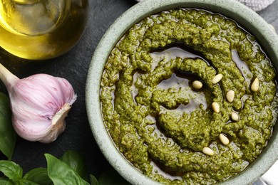 Photo of Tasty pesto sauce in bowl, basil, garlic and oil on black table, top view