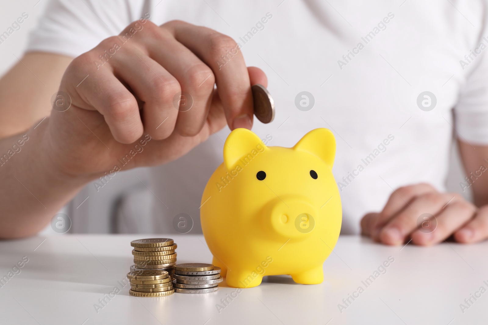 Photo of Man putting coin into yellow piggy bank at white table, closeup