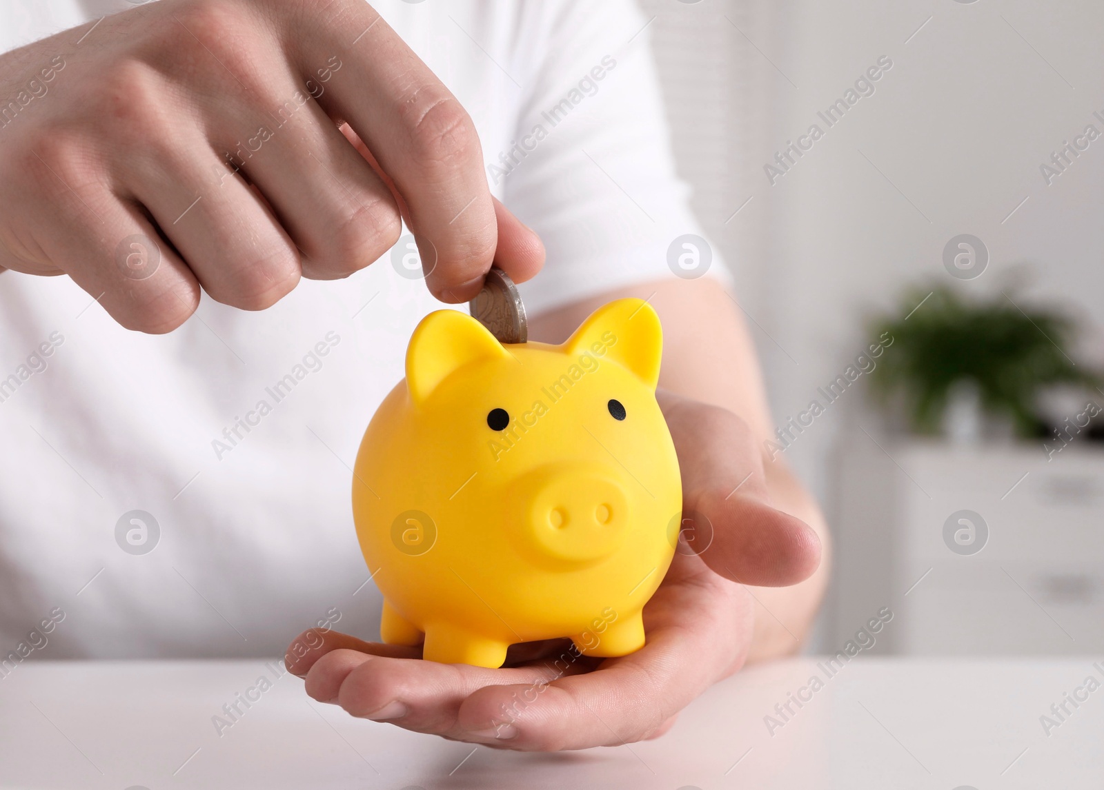 Photo of Man putting coin into yellow piggy bank at white table, closeup