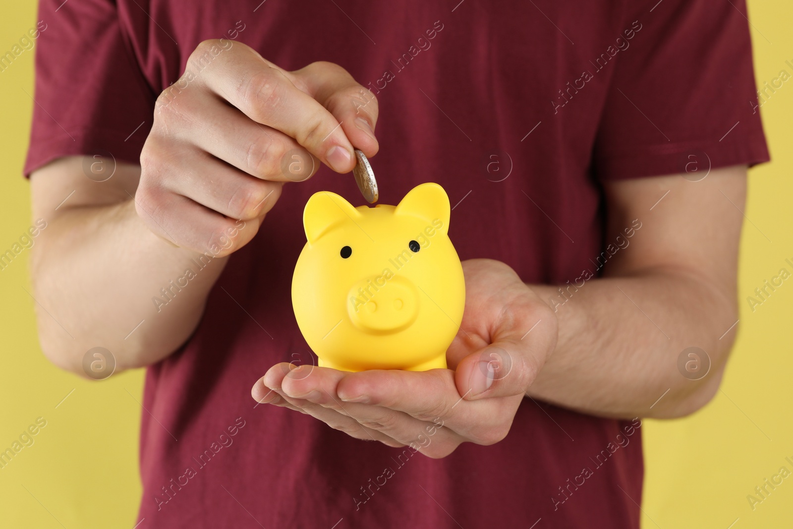 Photo of Man putting coin into piggy bank on yellow background, closeup