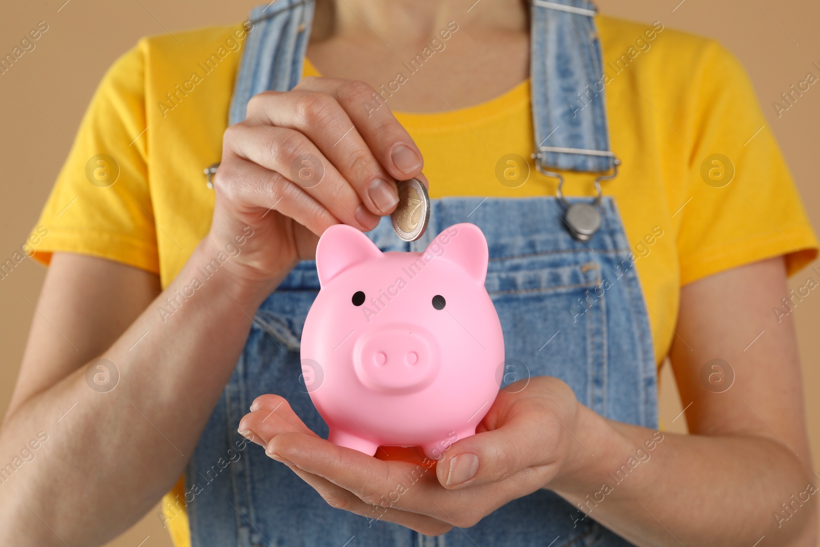 Photo of Woman putting coin into pink piggy bank on light brown background, closeup