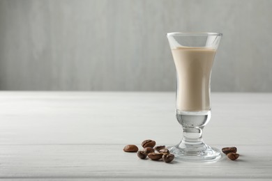 Photo of Coffee cream liqueur in glass and beans on white wooden table, space for text