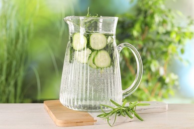 Photo of Refreshing cucumber water with rosemary in jug on light wooden table against blurred green background