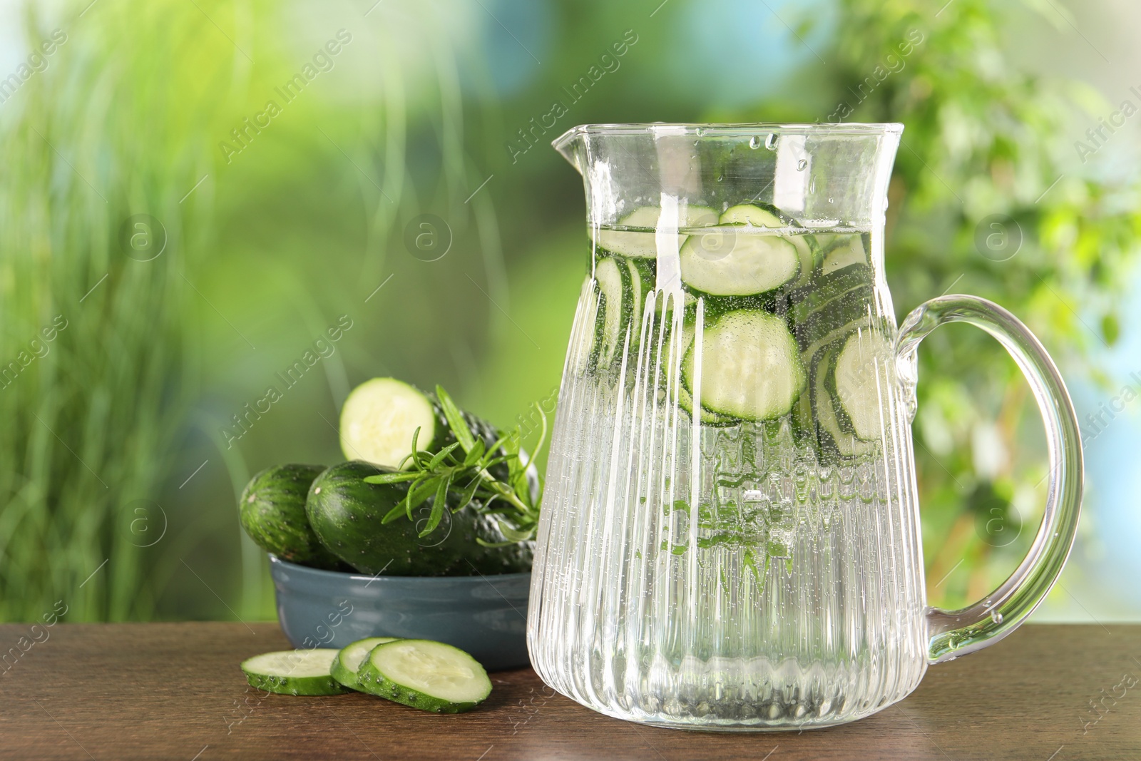 Photo of Refreshing cucumber water with rosemary in jug and vegetable on wooden table against blurred green background. Space for text