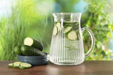 Photo of Refreshing cucumber water with rosemary in jug and vegetable on wooden table against blurred green background