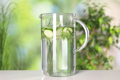 Refreshing cucumber water with rosemary in jug on light table against blurred green background