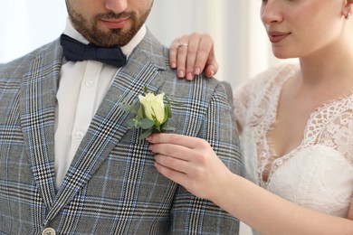 Photo of Bride putting boutonniere on her groom's jacket against light background, closeup. Wedding accessory